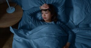 Woman lying in bed under blue sheets, arm resting on her forehead, looking tired or contemplative in dim light.