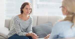 Woman in a striped long-sleeve top sits on a sofa, talking calmly with a therapist in a bright, calm office.