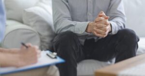 Man sits on a sofa with hands clasped in front of him during a counseling session, as a therapist takes notes on a clipboard nearby.