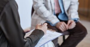 Clinician writing on a clipboard while a patient sits nearby awaiting consultation requesting notes.