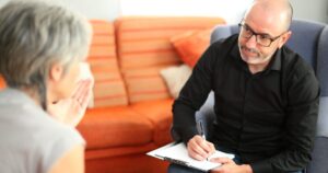 Counselor wearing glasses writes on a clipboard during a therapy session with a seated client in a cozy living room.