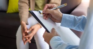 Close-up of a person writing on a clipboard held by another person during a consultation or interview on a couch.