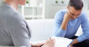 Therapist taking notes on a clipboard while a client sits beside, deep in thought during a counseling session.