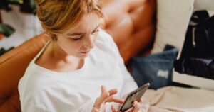 Woman in a white top sits in bed, looking at and tapping a smartphone in her hands.