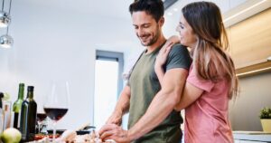 Couple cooking together at a kitchen island, smiling as they prep meat with wine on the counter.