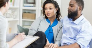 Couple sits on a sofa talking to a counselor who notes on a clipboard nearby.