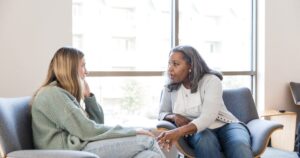 - Treat Mental Health Tennessee Two women sit on lounge chairs by a large window, having a thoughtful conversation in a bright room.