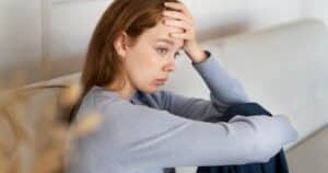 - Treat Mental Health Tennessee Young woman sits on a couch with her hand on her forehead, looking sad and thoughtful.