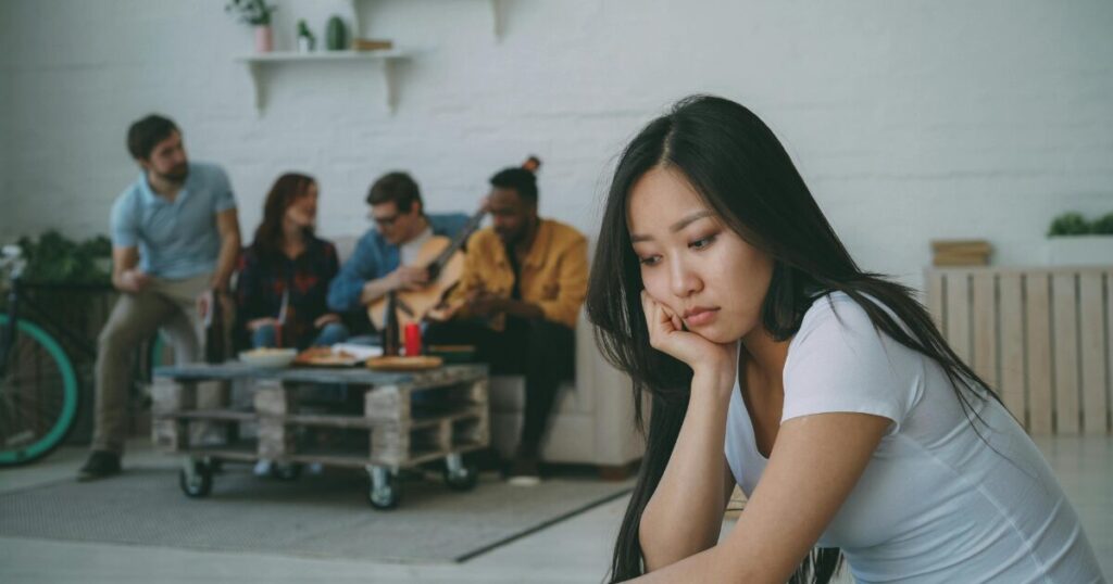 Woman in the foreground looks sad and distant, resting her chin on her hand while a group of friends chat and play guitar in the background.