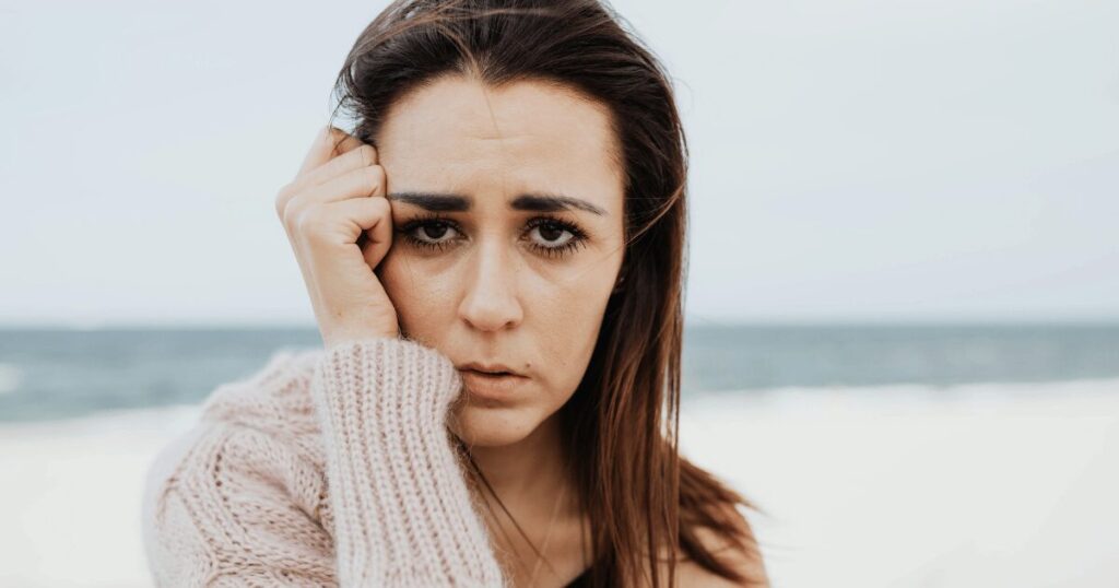 Close-up of a woman at the beach with a worried expression, touching her temple with her pink knit sleeve.