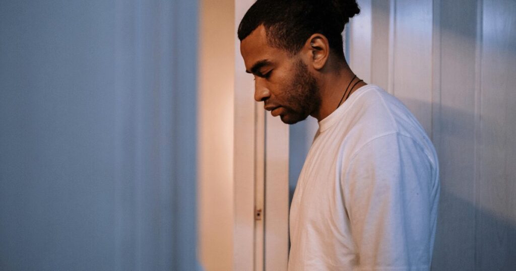 Young man in a white shirt stands profile against a doorway, looking down with a contemplative expression.