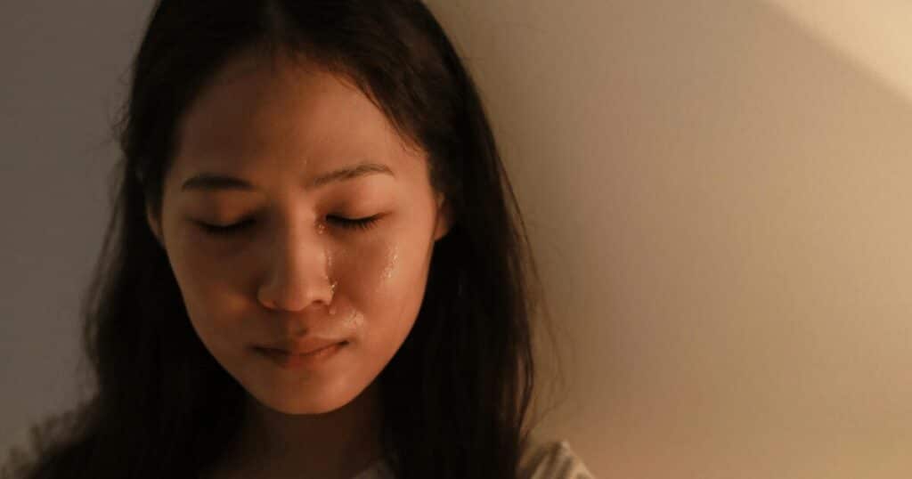 Close-up of a young woman with her eyes closed and a single tear running down her cheek under warm light.