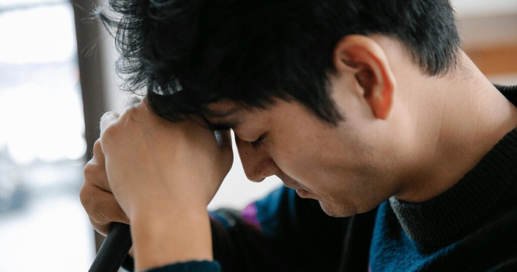 Man sitting indoors, head bowed and resting on his folded arms, appears stressed or tired by a window.