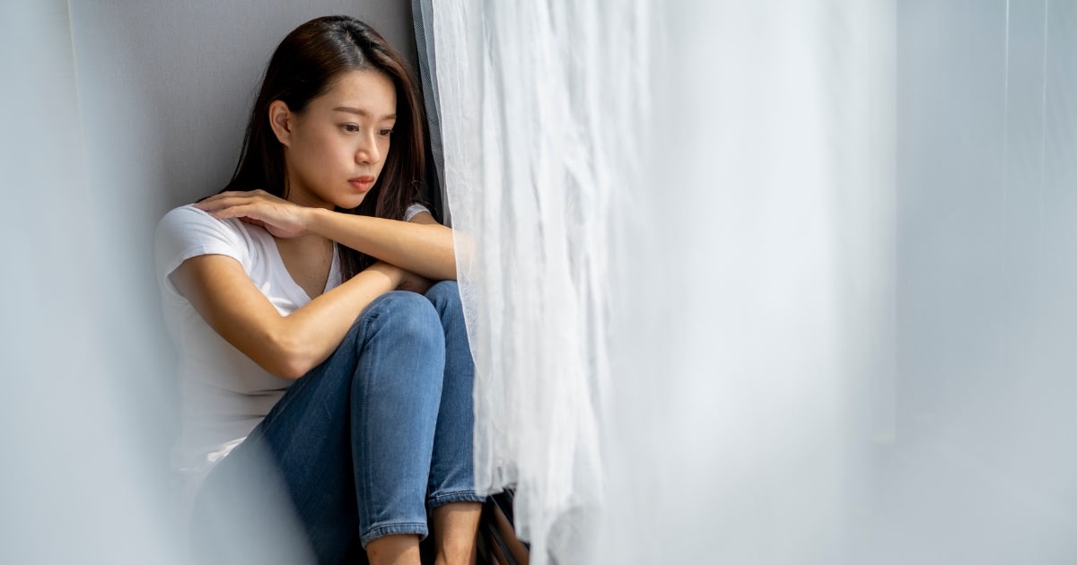 Young woman sitting in a corner with knees drawn up, arms resting on her knees, looking down with a sad expression.