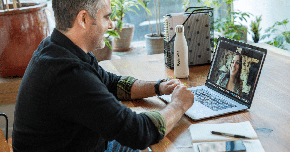 Man in a black shirt sits at a wooden desk, video calling a woman on his laptop screen, with a water bottle and notebook nearby.