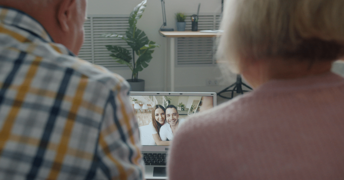 Two people with their backs to the camera watch a video call on a laptop, showing a smiling couple on screen.