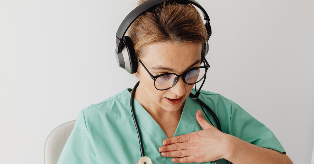 Healthcare professional in teal scrubs with stethoscope and headset, hand resting on chest as if listening or showing empathy.