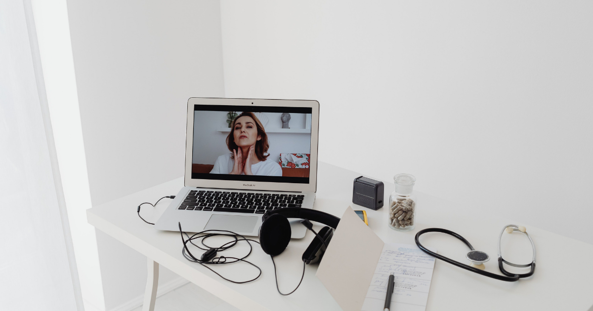 Telemedicine setup on a white desk: laptop on screen shows a woman during a video call, with headphones, stethoscope, and notes nearby.