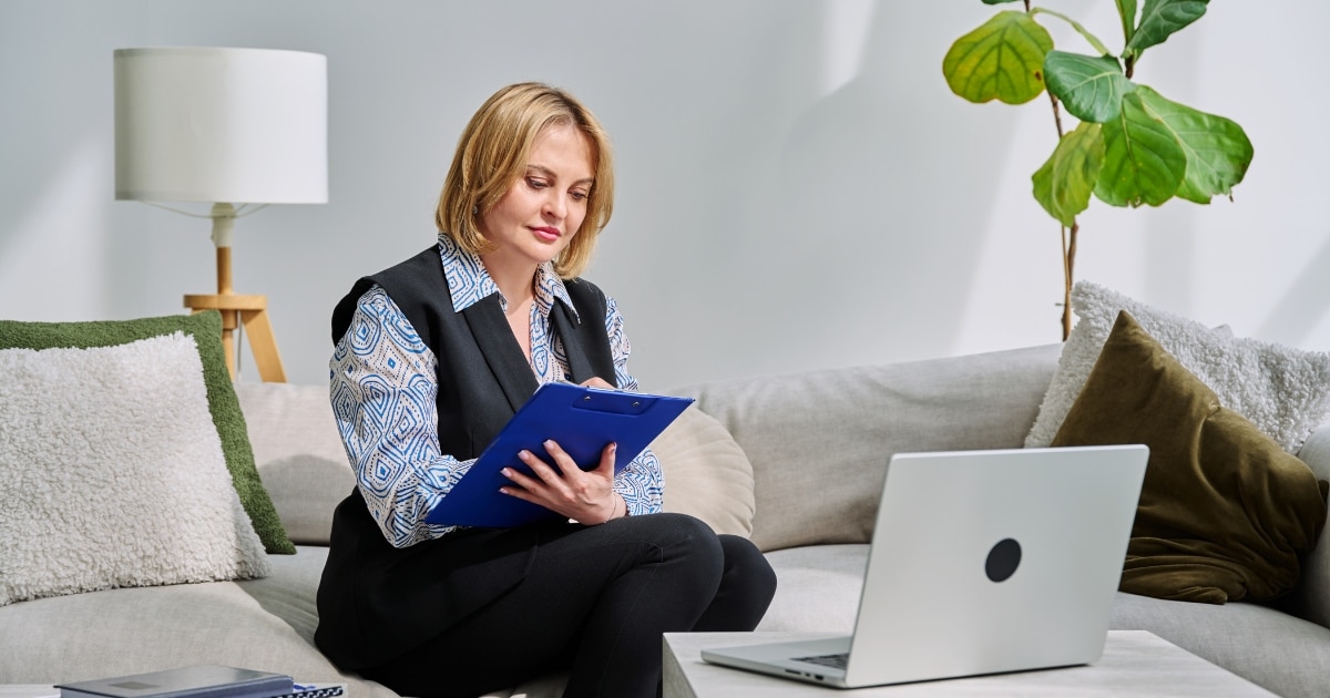 Professional woman sits on a sofa, reviewing notes on a blue clipboard with a laptop nearby on a coffee table.