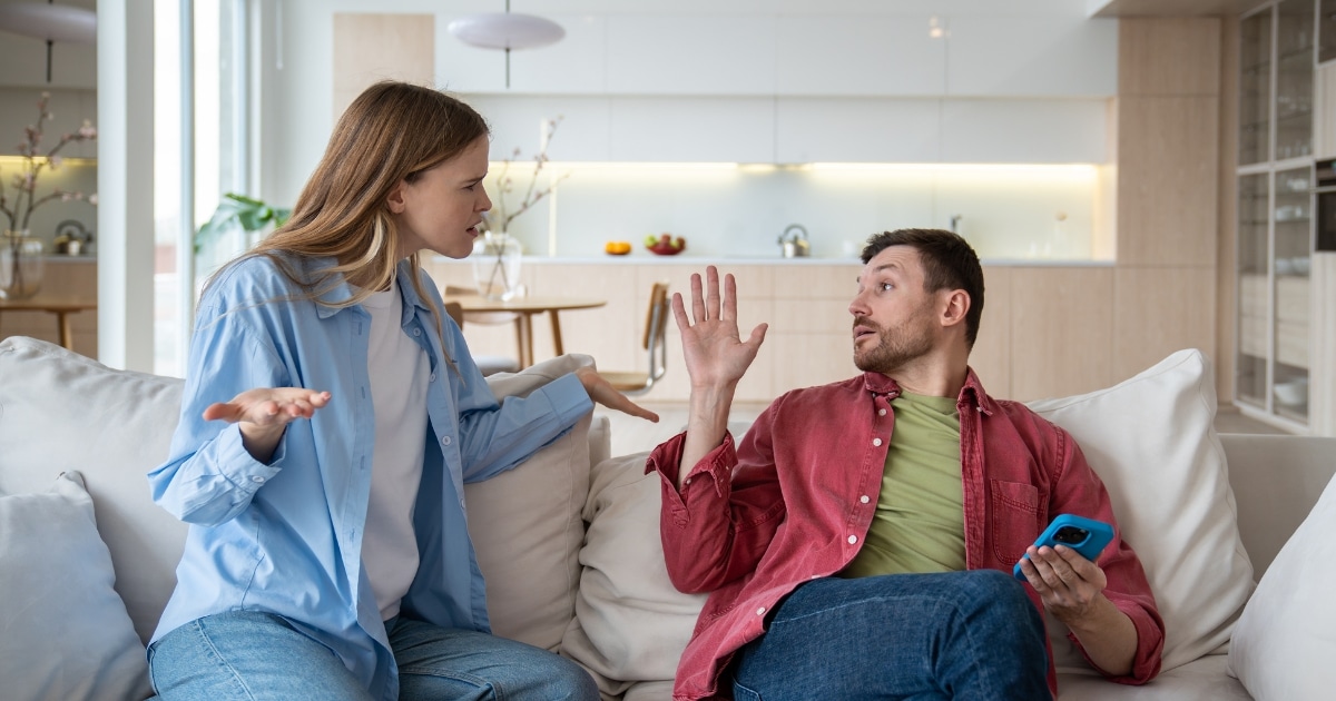 Woman on a sofa gesturing with open hands as she argues with a man who holds a blue smartphone. (Bright, modern living area in background)