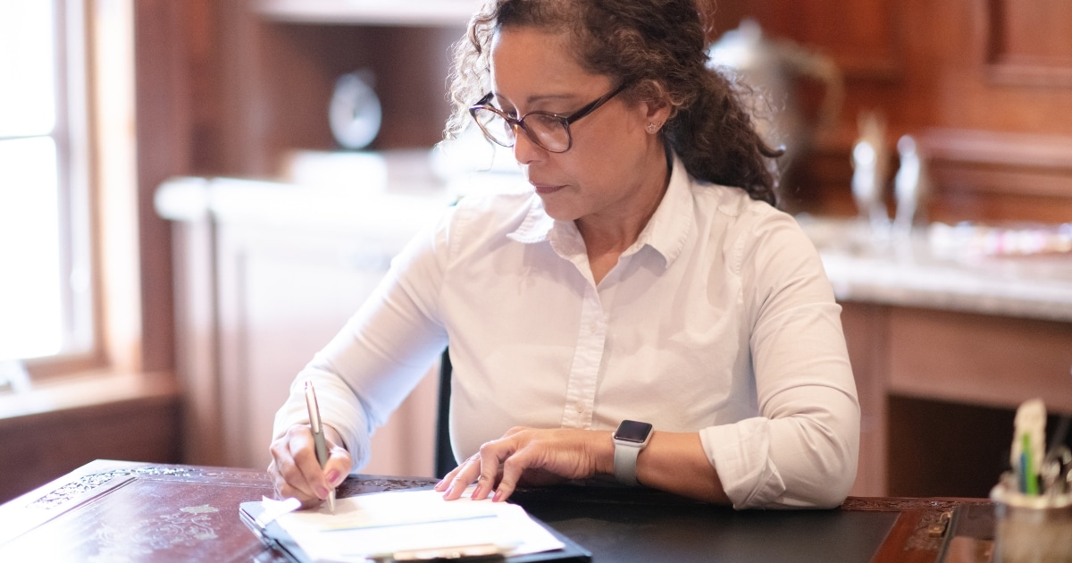 Woman in a white button-down shirt and glasses writes on a notebook at a wooden desk in a home kitchen, wearing a smartwatch on her left wrist.