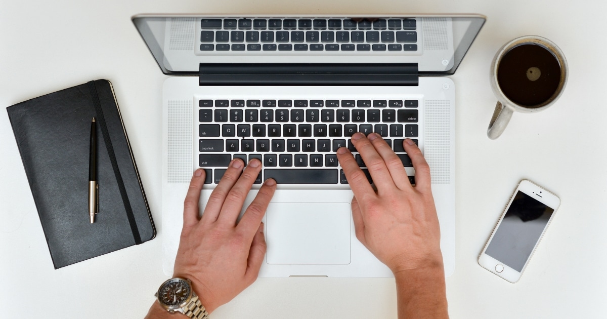Hands typing on a laptop keyboard on a white desk, with a notebook, pen, coffee mug, and smartphone nearby.