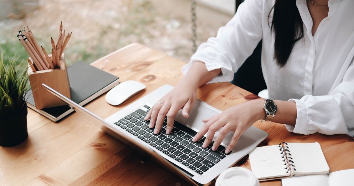 Person typing on a silver laptop at a wooden desk, with a notebook, pencils, plant, and a coffee cup nearby.