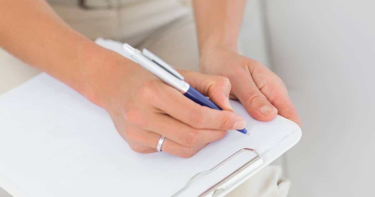 Close-up of hands writing on a clipboard with a blue ballpoint pen on white paper
