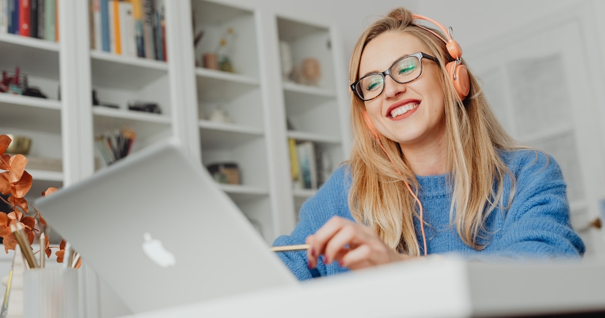 Smiling woman with glasses and pink headphones using a silver laptop at a white desk in a bright room.