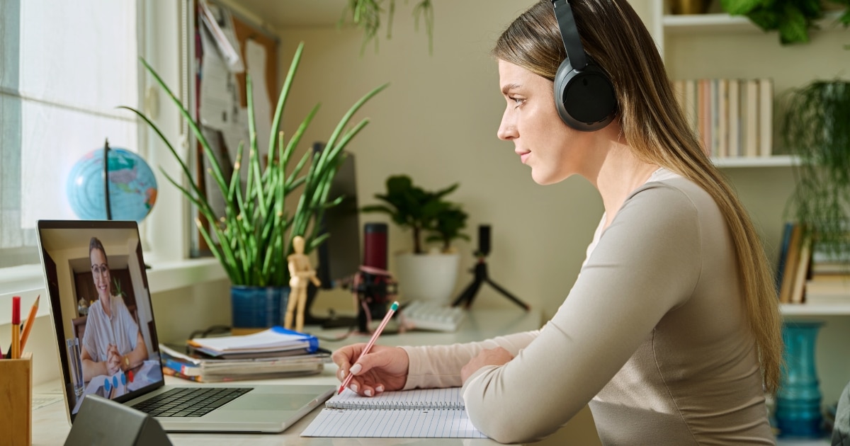 Woman wearing headphones, watching a video call on a laptop while taking notes in a notebook at a tidy desk.