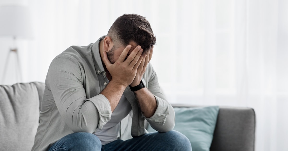 Man sitting on a couch with his face buried in his hands, looking stressed or overwhelmed.