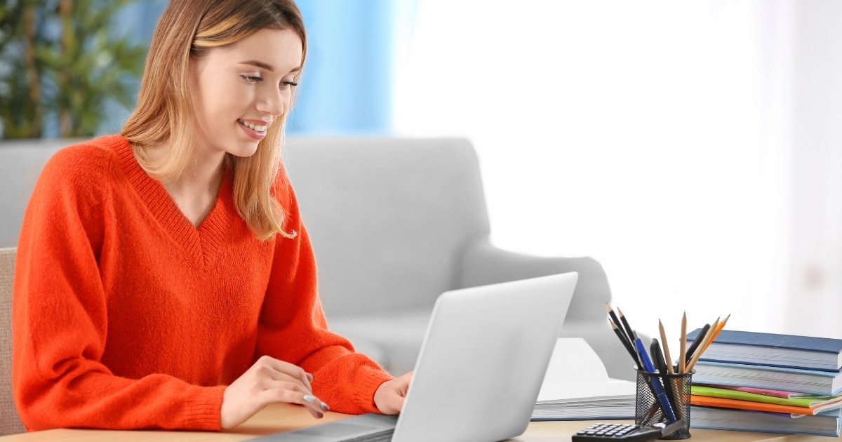 Woman in an orange sweater typing on a laptop at a desk, with pencils and books nearby.